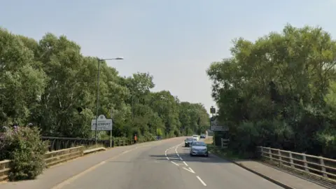 Google A black road, with a broken white line in the middle, edged by green bushes. On the left is a white rectangular sign with the words "Aylesbury" written in black capitals. On the right is a line of approaching cars.