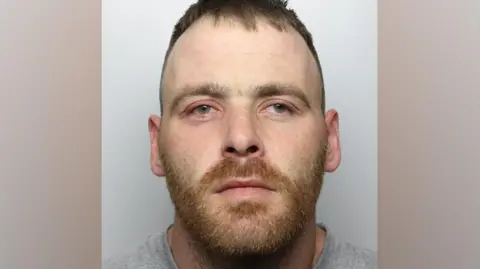 West Yorkshire Police A custody image of Mark Ross. He has a short red beard and is looking away from the camera. 