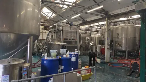 A room in a brewery, with many large silver tanks visible.