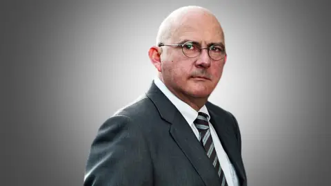 The Times A portrait of Andrew Norfolk wearing a suit and tie with glasses on looking directly at the camera against a grey background.