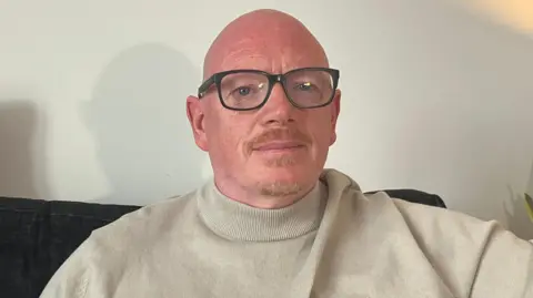 A man with glasses in a beige turtleneck sits on a sofa. 