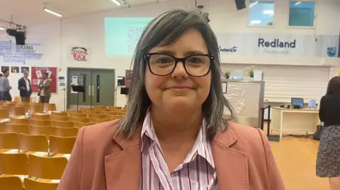 Melanie Sweet standing in a school hall. She has medium length straight, grey hair and is wearing thick-rimmed black glasses, a pink and white striped shirt and a salmon pink blazer.