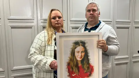 Shows a woman with blond hair to the left, a man in grey hair and light-coloured hoodie to the right. They are holding a framed photograph of a teenage girl with long hair, with sunglasses on the top of her head. She is wearing a Liverpool jersey. 