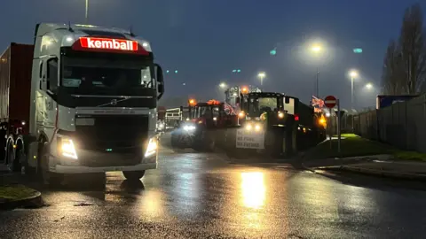 Darren Rozier/BBC Tractors decorated with Union Flags and protest signs are parked over a junction that enters a port, a lorry is parked on the other side of the road