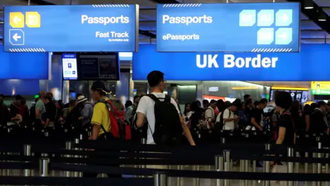 People queue at passport control at the UK border. Signs overhead read 'Passports Fast Track', 'Passports ePassports' and 'UK Border'. 