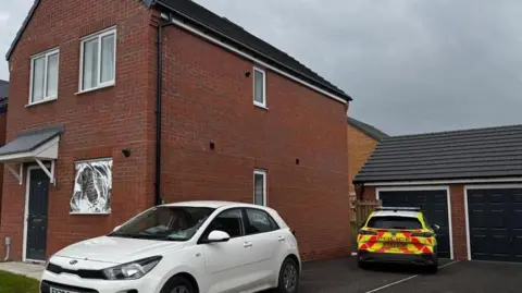 Two police vehicles are parked on the drive of a newbuild house. One is a white car without any police markings on. A police officer is sat in the driver's seat. The other car has full police colours. There is a silver foil sheet over the downstairs window of the house.