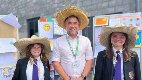Jeorgie, 11, (left), Lee Thomas (centre), and Izzy, 11, (right) are stood wearing straw hats. They are smiling stood in from of sun safety posters made by students.