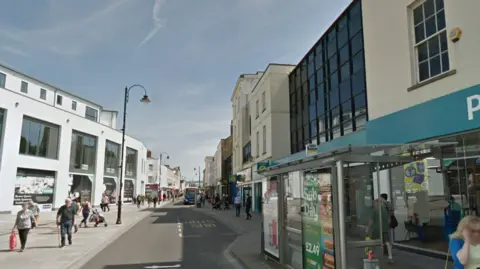 Street view of High St in Cheltenham. The edge of Poundland is visible to the right. A bus has pulled into a stop in the distance, and the street shows lots of people walking up and down.