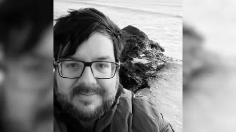 Matt Winkworth A black and white selfie of a bearded man wearing glasses, with the sea and some rocks behind him.