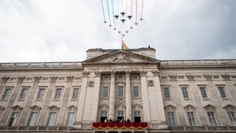 PA Media Red Arrow planes and fighter jets fly over Buckingham Palace. The Royal Family are on the balcony, which is draped with red cloth.