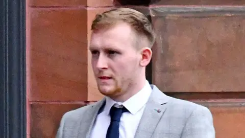 Alan Lewis Christopher Morrow pictured outside court, he has short brown hair and ginger stubble. He is wearing a light grey suit jacket, a white shirt and a dark blue skinny tie.