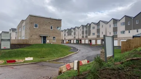 Daniel Mumby The housing estate in Minehead. There is a row of 12 modern terraced homes built on one side. They are three storeys high, with garages to the bottom floor. There is an unfinished road meandering to the left and some terraced houses opposite the row.