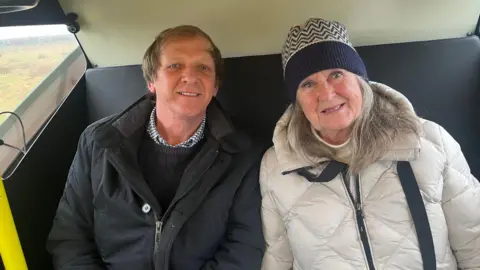 A man with a black jacket and a woman with a white jacket and blue and white hat sitting on a seat next to each other.