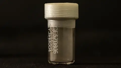 A small clear viall with a white plastic top and tape around the lid. It is lit against a black background. On the front is a code. Inside it looks great and at the bottom there is a coating of dust.
