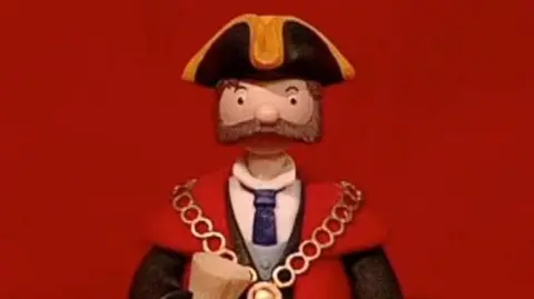 The Mayor from the stop motion children's show, Trumpton. He has a large brown beard and is wearing a black and yellow tricorn hat, a red and black robe and a gold chain. He is standing against a red background