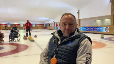 Paul Simmons wears a dark blue gilet over a fleece zipped top. He has grey short hair and a trimmed beard.