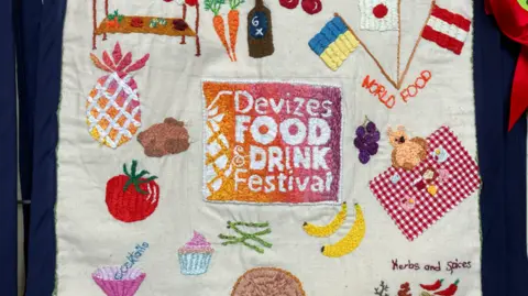A fabric banner square with embroidery on it depicting food and drinks - including a tomato, a pineapple, bananas and a cupcake. The middle of the banner reads 'Deivzes Food and Drink Festival'.