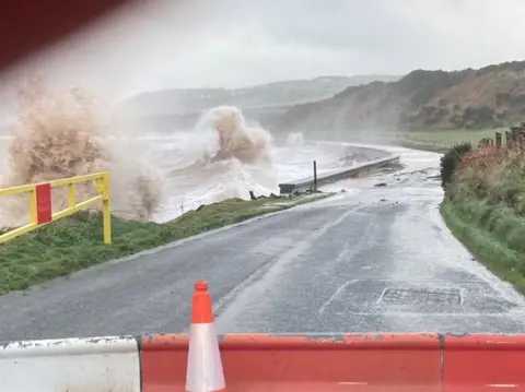 Dumfries and Galloway Council A coastal road with a wave crashing over the wall at Drummore in Dumfries and Galloway