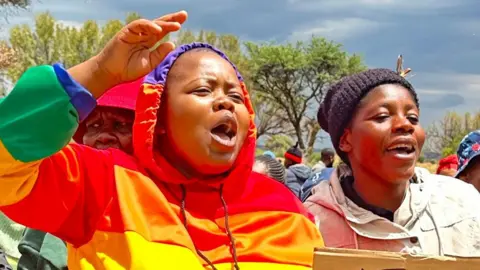 A group of female protesters, with one dressed in a rainbow hoodie, are shouting.