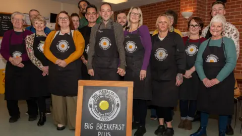 About a dozen volunteers all wearing Big Breakfast Plus branded black aprons stand behind a small chalk board with the Big Breakfast Plus logo on it