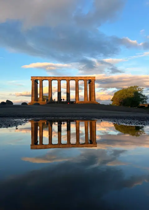 Leora Soibelman Monument with tall columns silhouetted against a partly cloudy sky at sunset, reflected in a puddle in the foreground.
