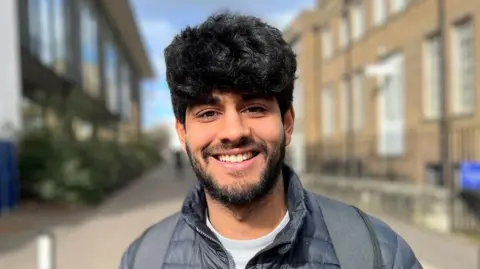 A man with dark hair and facial hair smiles at the camera. He wears a navy jacket and grey backpack. A street scene is blurred behind him and it is a bright sunny day.