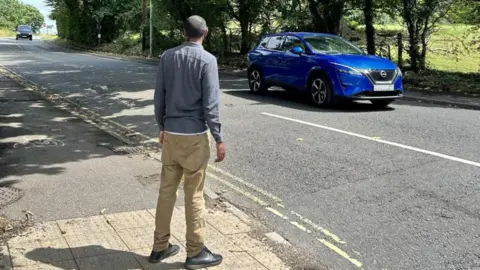 Dorset Council A man wear brown trousers and a blue shirt trying to cross a busy A road