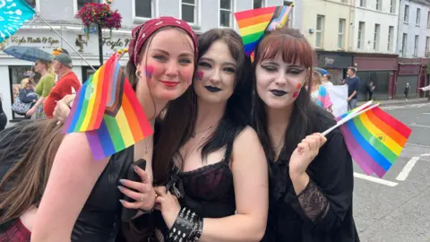 Three people smiling at the camera. All three of them are holding pride flags, and have LGBT+ flags painted on their faces. 