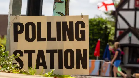 A sign says 'Polling Station.' In a faded out background, a person can be seen walking and there is an England flag on a Tudor style building.