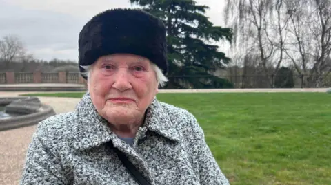 Kate Bradbrook/BBC Peggy Lade, a woman who is outside in front of a grassy lawn. She is looking directly at the camera and is wearing a black and white coat and black fur hat.