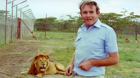 Hugh Cran in the foreground with a lion behind in an animal enclosure