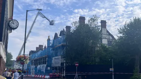 A black and white building covered in scaffolding with blue netting around it. There is a white fire service aerial platform with a firefighter spraying a hose of water on the roof. The area is cordoned off with red and white tape