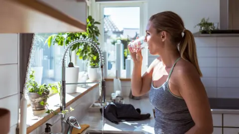 A woman in a vest top with blonde hair tied at the back drinks a glass of water standing by a kitchen sink. The white-tiled kitchen has pots of green plants on the window sill and has a big glass window allowing sunlight into the kitchen. 