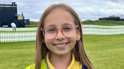 A girl wearing glasses and a yellow polo shirt looks in the camera. In the background is a grassy lawn, like the fairway of a golf course, and a white wooden fence. 