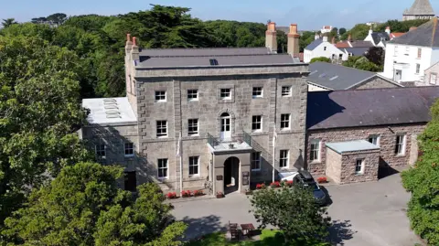 A drone shot of Alderney Island Hall. It is a grey-stone Georgian building with long windows and a large porch area. The building is surrounded by trees.