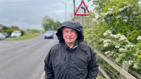 A man in a black rain jacket with the hood pulled up stands next to a roadway with a sign for a dangerous bend above his head.