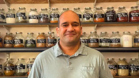 BBC/ Emily Johnson A man stands in front of neatly arranged wooden shelves filled with glass jars containing spices and dried goods. Each jar is labelled with the item name and price per 10 grams, written in white chalk on black tags.