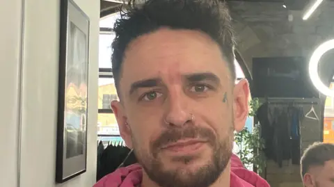 A man with short brown hair wearing a pink hoodie stands in a barber's shop. He has a tattoo on his left cheek and two nose piercings.