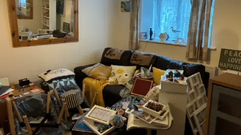 A wide image of the mess in David's living room. A black leather sofa is covered in cushions and blankets. In front, is a pile of items, which include upside down chairs, photo frames and ornaments.