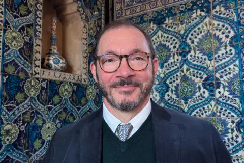 BBC/ Luxmy Gopal A man with close cropped dark hair and beard looks at the camera. He is bespectacled. He is wearing a dark jacket and a shirt and tie