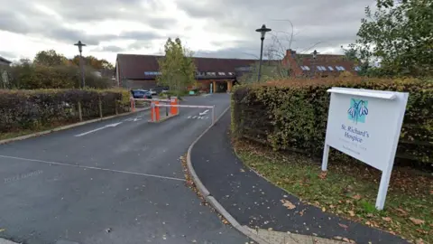 Google The entrance to St Richard's Hospice, showing a sign from the roadside and two car park barriers