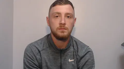 GMP A police mugshot showing a man in his 20s, with light brown hair and ginger goatee style facial hair, looking into the camera with a blank expression. He is wearing a grey zip-up Nike tracksuit top. 