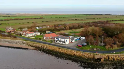 Chris @apsdrones An aerial view of coastline in Kilnsea showing a white one-storey pub in the middle of the photo, which is close to the cliff-edge. A few houses can be seen dotted around it, as well as cars parked up, caravans, and then several fields in the distance.