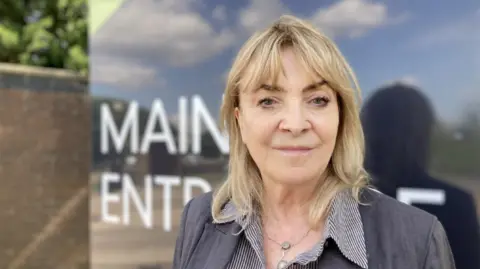 A woman with blonde shoulder-length hair is looking directly at the camera, with a closed smile. She is wearing a grey jacket and striped blouse.