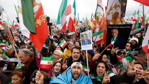 AFP via Getty Images A sea of protesters are stood together to denounce the Iranian government. Many of them are carrying the Iranian flag and posters of Reza Pahlavi, who is a political activist and Iranian dissident in exile. The sky is grey behind them.