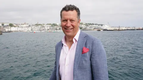 Bank Aston Jay Goss is pictured smiling at the camera, against the backdrop of the sea. He is wearing a smart, grey suit jacket, with pale pink shirt underneath. He has brown hair and is pictured amidst the backdrop of the sea, with a boat and housing in the background.