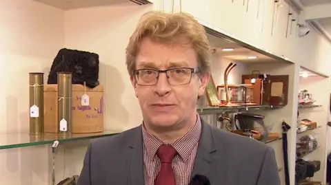 Brian Goodison-Blanks looking at the camera. He is wearing glasses, a grey suit and red and white checked shirt, with a red tie.