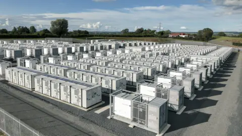 A wide aerial view of a large energy storage facility containing hundreds of white, container-sized battery units arranged in neat rows on a gravel site. The installation is surrounded by fencing, grassy fields, and scattered trees under a partly cloudy sky. Rural buildings are visible in the distance.