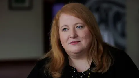 Liam McBurney/PA Media Naomi Long, a woman with shoulder-length ginger hair, looks beyond the camera in a photoshoot.  She is wearing a black cardigan over a black top or dress with white polka dots and a yellow floral pattern. 
