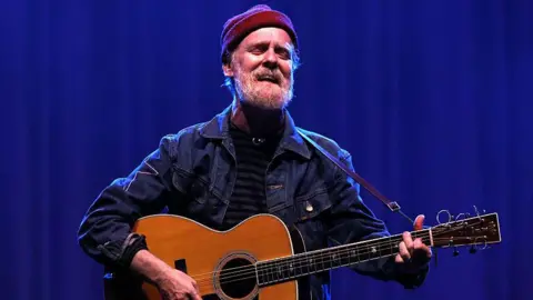 Getty Images Glen Hansard sings while holding an acoustic guitar. He has a grey beard and is wearing a red wool hat and denim jacket. He is standing in front of a dark blue curtain.